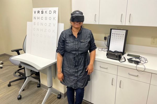 Sarika wearing an AR headset, standing in front of a table with low vision tools on it, smiling.