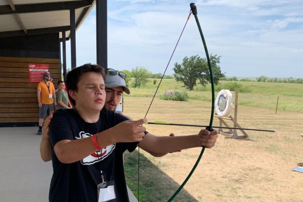 A visually impaired boy holding a bow and arrow and being shown how to aim the arrow.