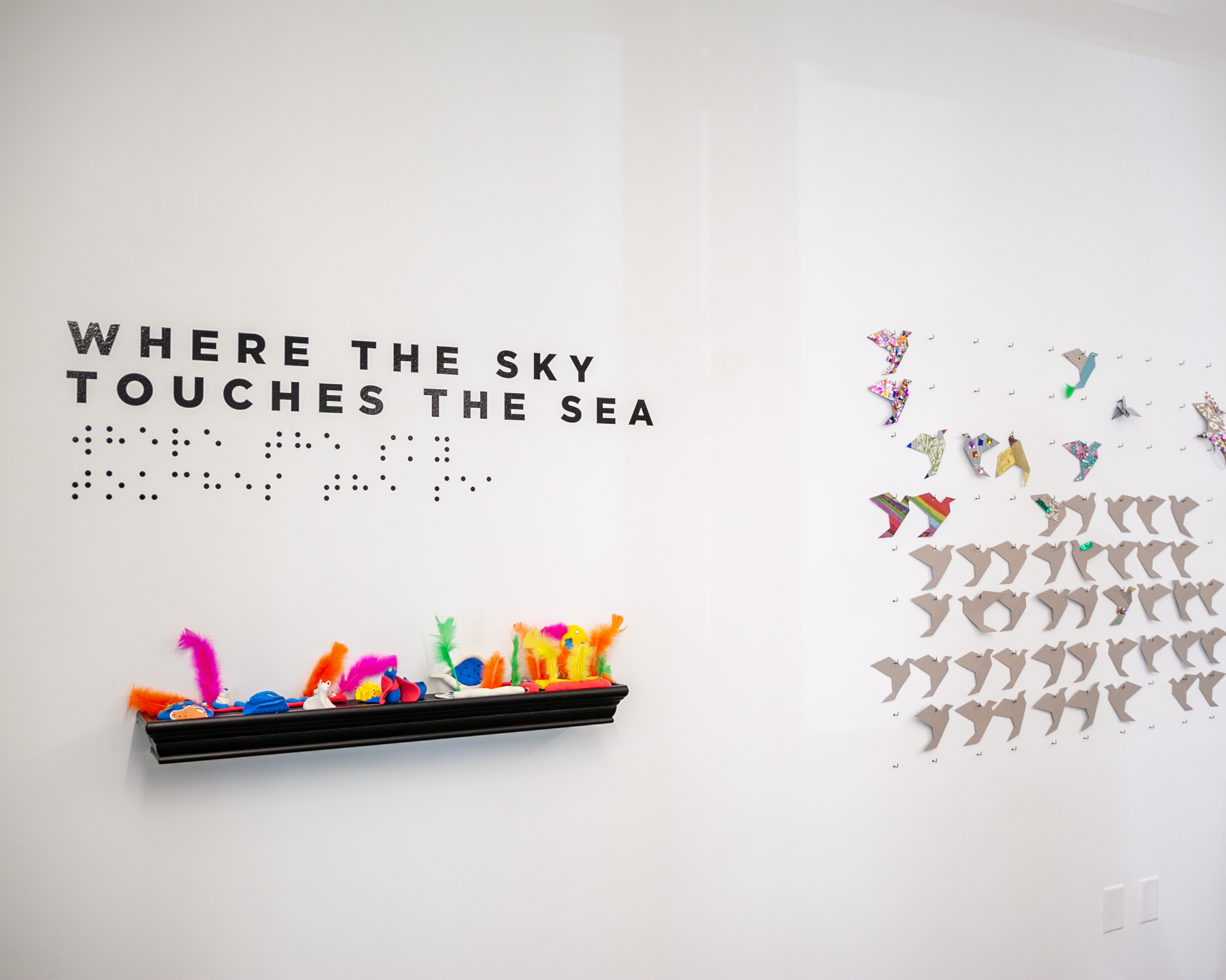 Envision Arts Gallery wall with text that says where the sky touches the sea in big and braille below and origami shapes on the other wall