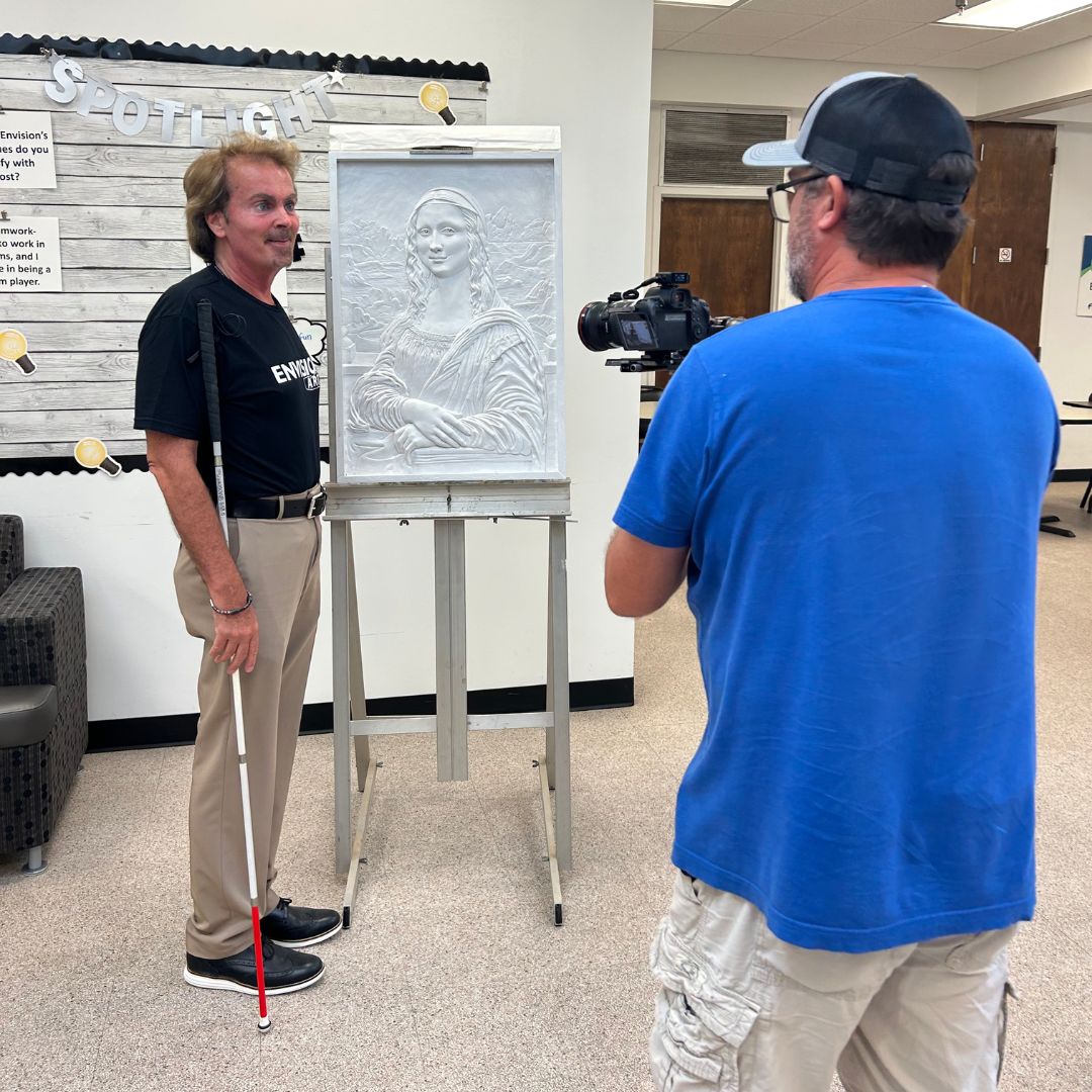 Blake Lindsay standing next to a piece of Tomas Bustos' art on an easle as he speaks to a cameraman holding a camera and filming.