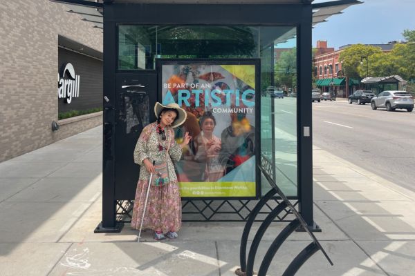 A wide angle shot of Tomiyo standing by her bus stop art and smiling at the camera.