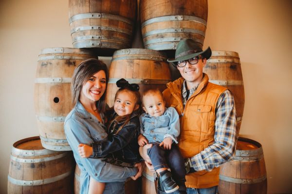 Dani and Dave standing in front of a wine barrel, both holding their two children, all smiling together.