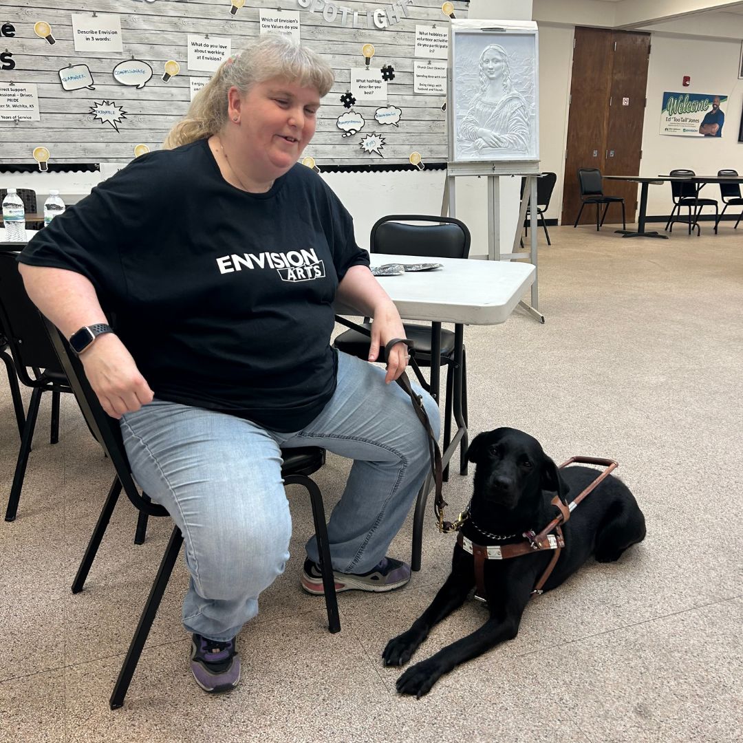 Keela Alonzo, arts participant, smiling for the camera as she sits in the art room next to her guide dog.