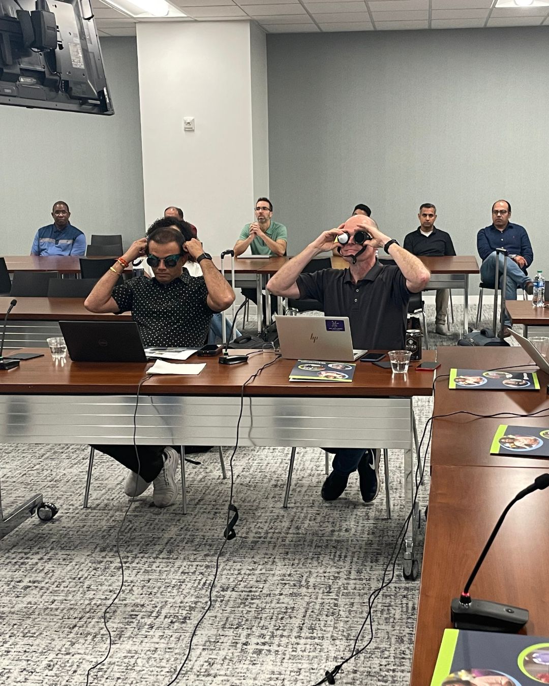Two sighted individuals sitting at a table trying on different goggles that simulate visual impairments.