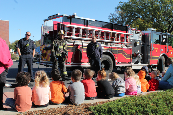 A group of about 15 children sitting outside while firefighters talk to them.
