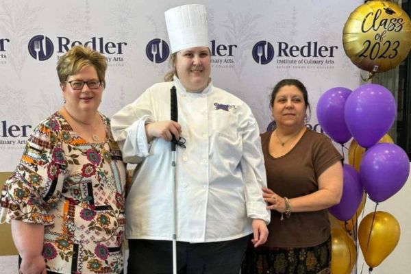 Lauren standing with two Butler community college professors, in her chef uniform and hat, smiling at the camera. Behind her are balloons that says "Class of 2023."