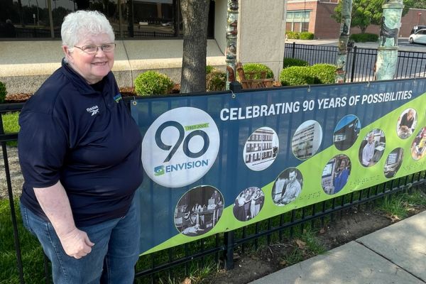 Wanda smiling as she stands next to the 90th banner at Envision.