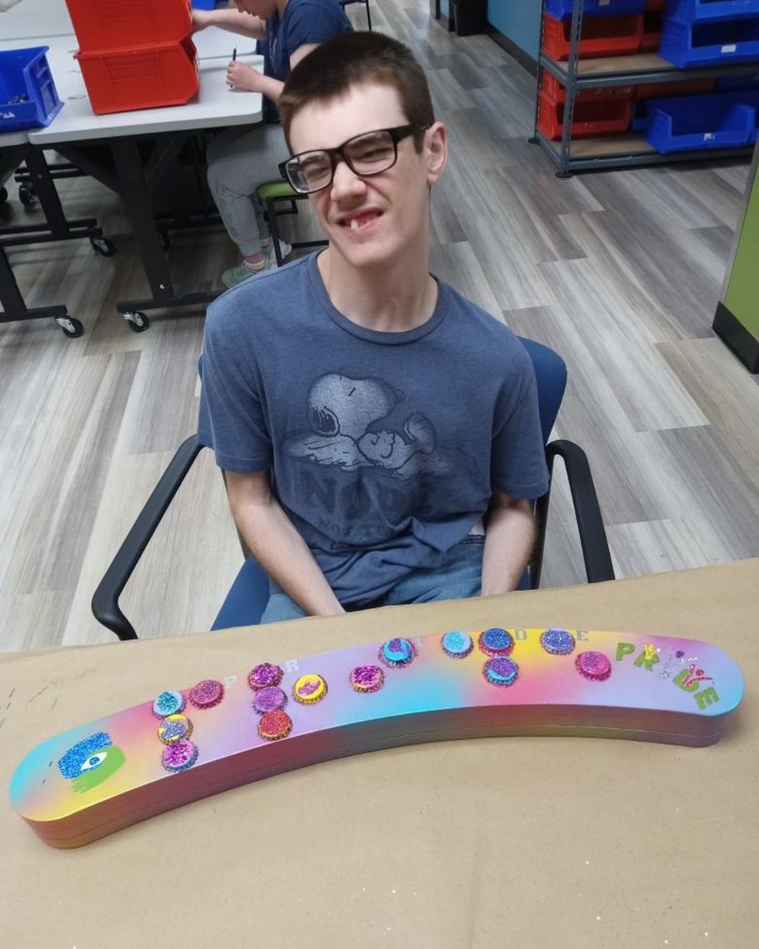 A young man sitting a table smiling as his artwork piece is in front of him.