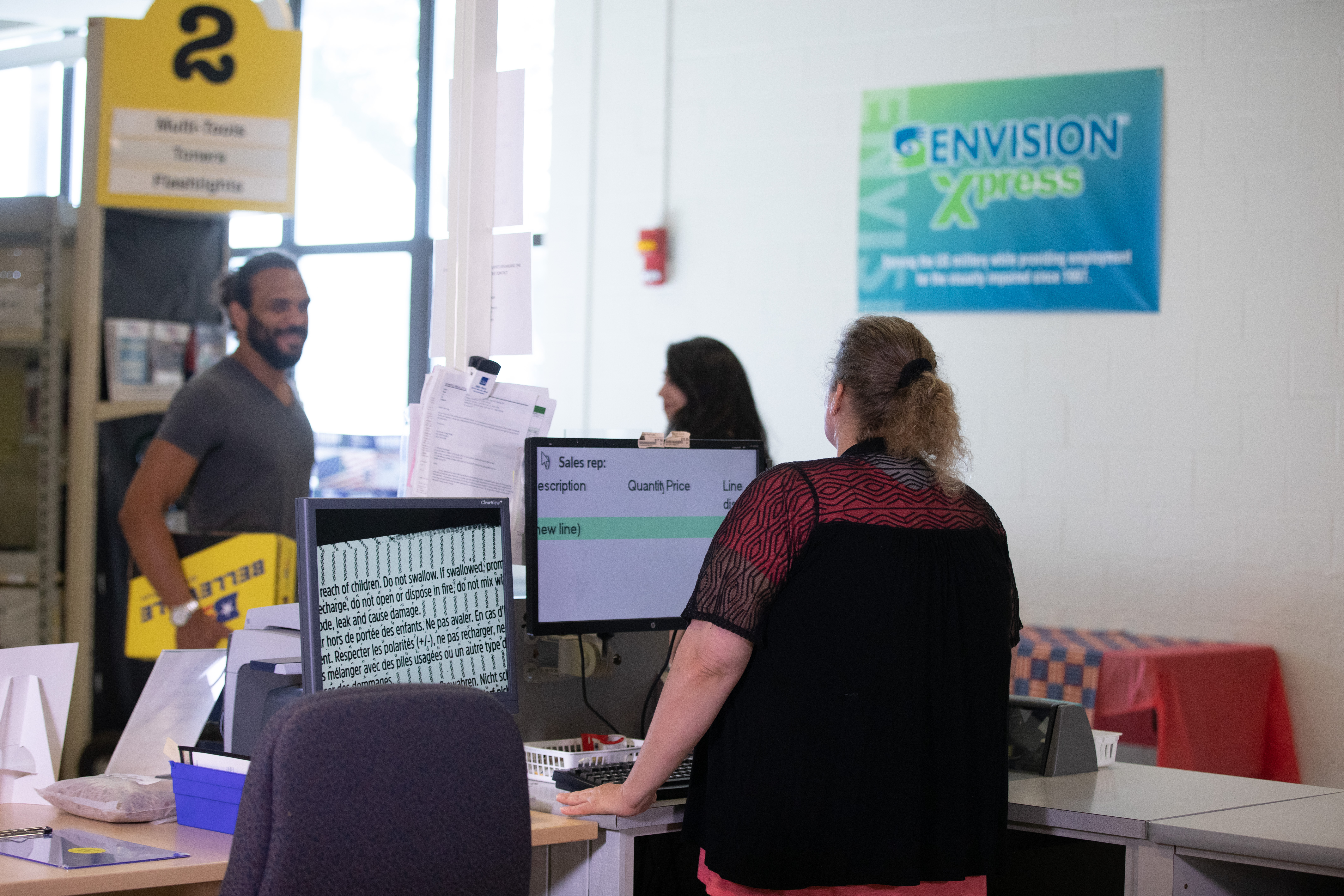 Envision Xpress sign on the wall, female cashier at computer helping male customer