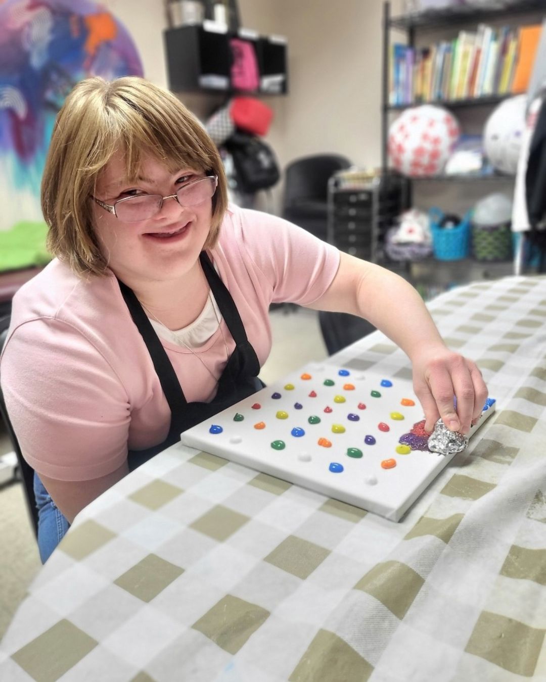 a young girl smiling at a table as she puts paint on a canvas.