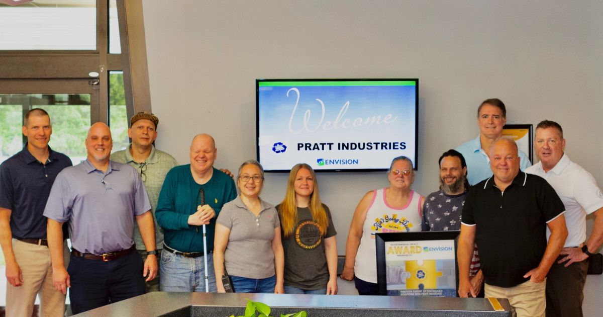 A group of Envision employees standing in front of a screen that reads "Pratt Industries," holding the award plaque and smiling.