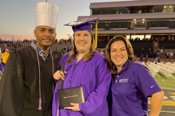 Lauren at graduation, standing with her two culinary teachers in her purple graduation cap and gown, with her degree and white cane in hand, smiling at the camera.