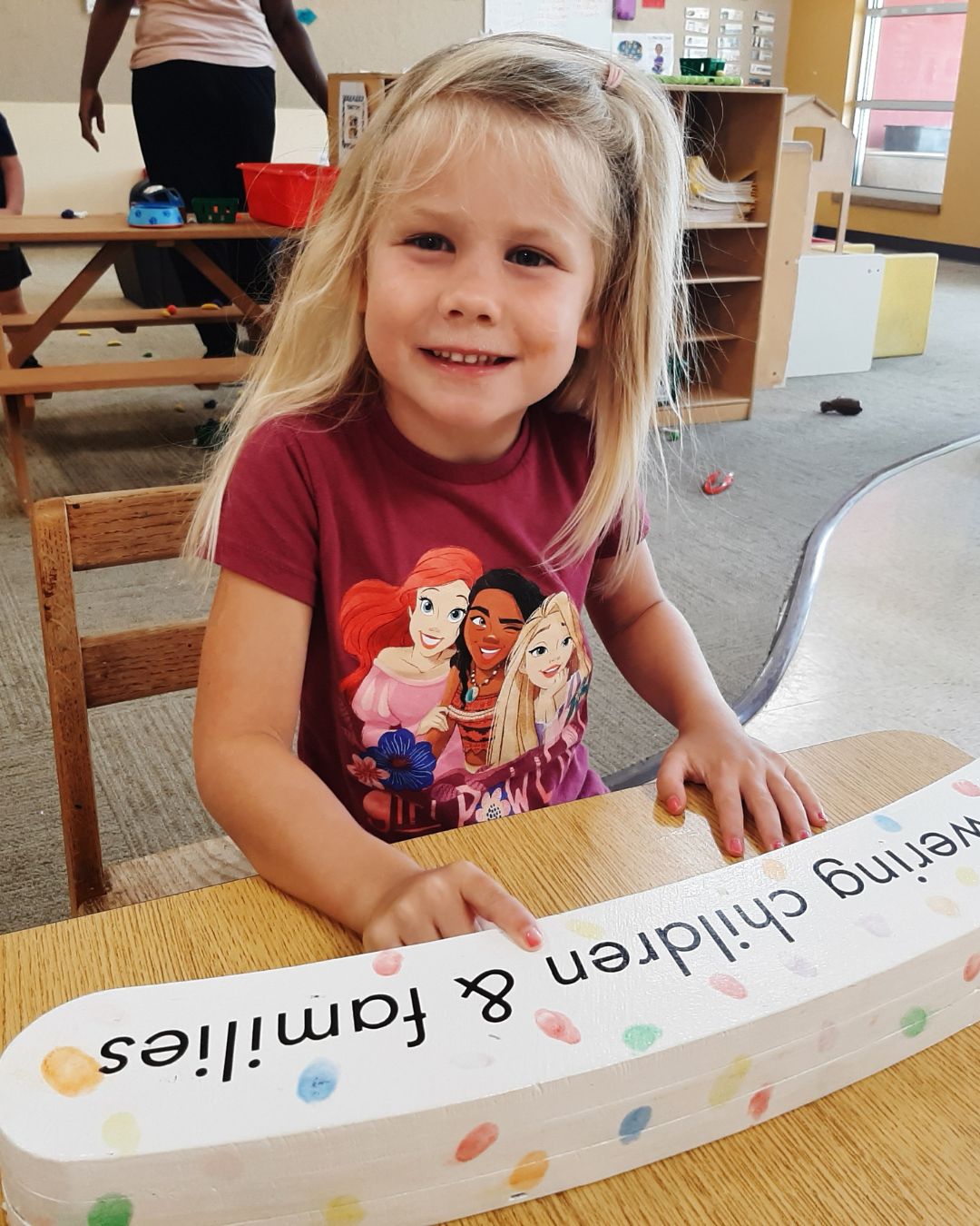 A young girl smiling at a table as she puts her hand on a piece of artwork.