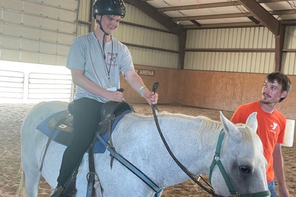 a visually impaired boy riding on a horse while a worker guides him out of the stable.