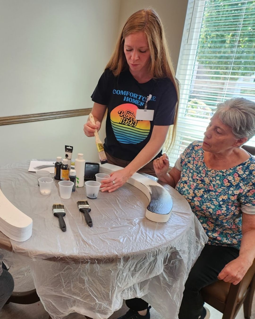 An older lady sitting at a table painting a piece of a canvas.