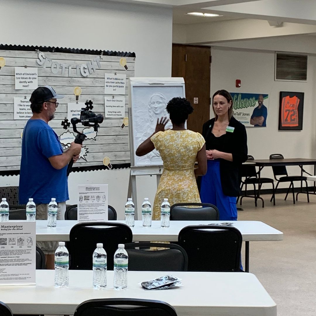 Sarah Kephart, Envision Arts Manager, standing next to a piece of artwork made by Tomas Bustos, speaking to an interviewer and a cameraman as he films.