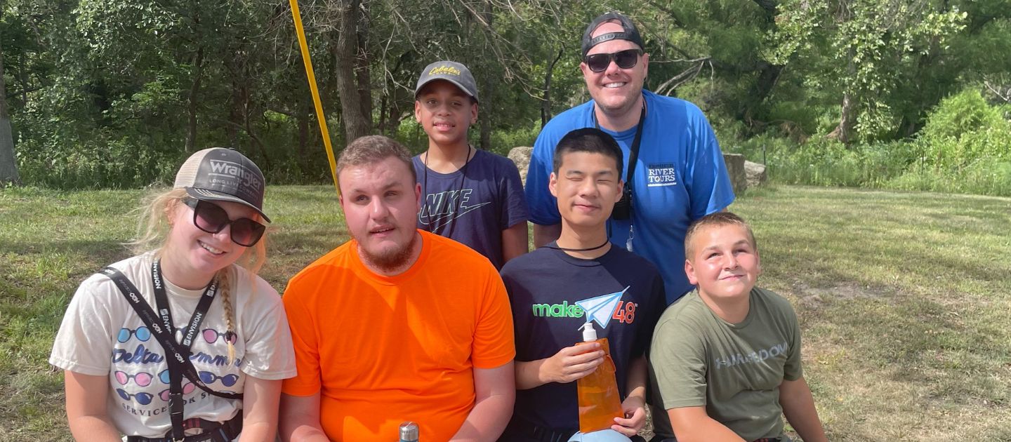 A group of 5 boys sitting on a bench smiling for the camera, as their camp counselor is talking to them.