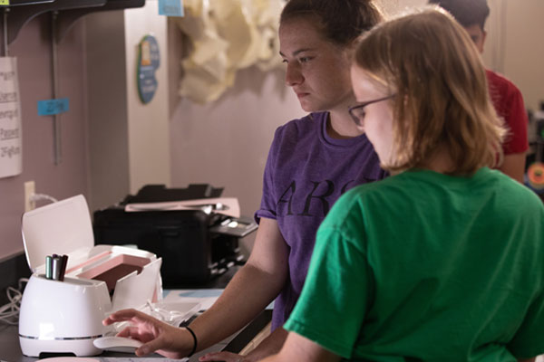 A volunteer helps an art participant set up a design on the laptop.