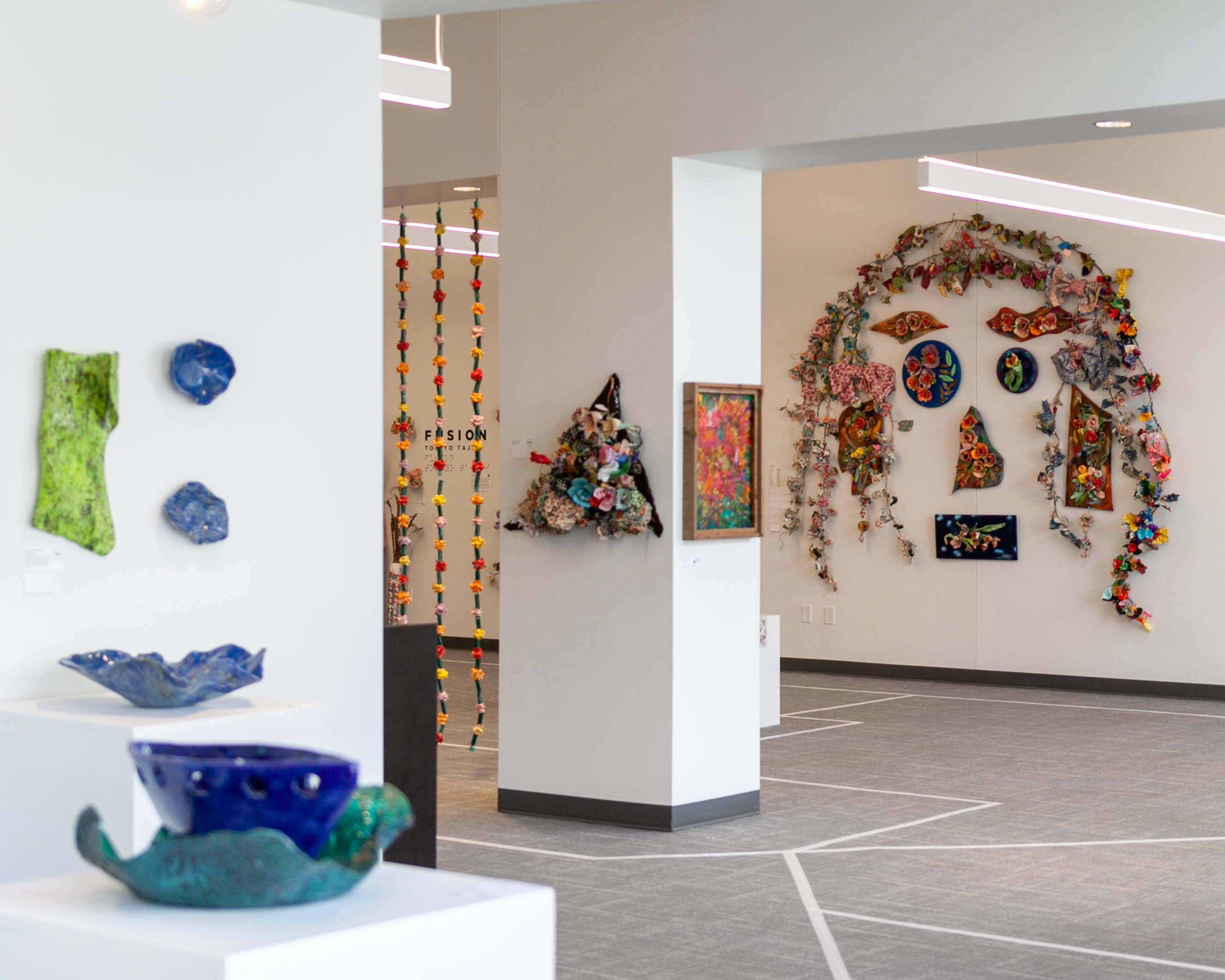 This image looks into the Envision Arts Gallery from the Patricia A. Window Gallery where Erica Johnson's exhibition Memory of Waves is on display. Several ceramic art pieces can be seen on pedestals and hung on the wall. In the background, Tomiyo Tajiri's exhibition titled Fusion can be seen