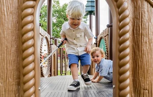 Two young boys playing on an inclusive outdoor playground. One boy, using a white cane, confidently explores the play structure while the other follows behind, highlighting accessibility and independence in play.