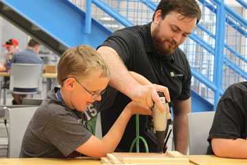 Young middle school student working on a piece of wood during a level up woodworking class while an adult helps him