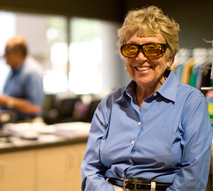 A cheerful elderly woman with short, wavy hair wearing large, eyeglasses and a blue button-up shirt. She is smiling broadly and appears to be seated, with what looks like an indoor setting blurred in the background.