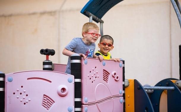 Two young boys smiling and playing on a pink and blue outdoor playground structure. Both boys wear glasses and appear to be enjoying their time together, engaging in imaginative play.