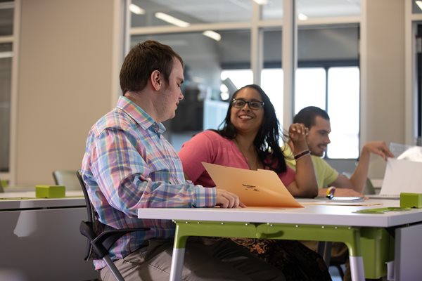 A woman in a coral shirt and glasses helping a man in a plaid shirt sitting at a desk. 