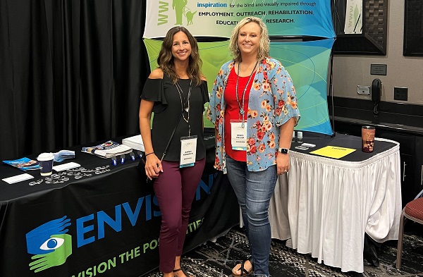 Two women posing together in front of an Envision table at a conference. 