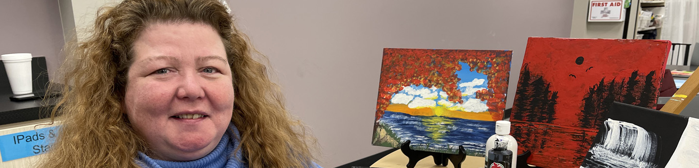 A woman in a blue shirt sitting at a wooden desk smiling for a photo with paintings behind her