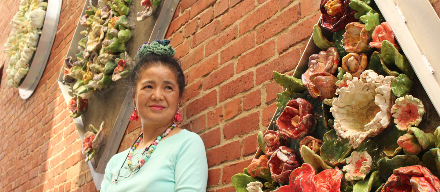 Tomiyo stands in front of a brick wall in Gallery Alley in front of her geometric and colorful ceramic flower art piece