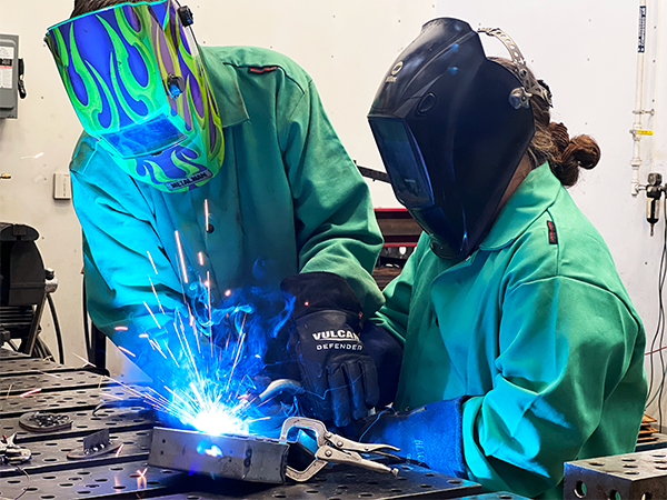 A BVI envision level up student with a mentor/teacher doing welding at WSU Go Create. Blue and orange sparks are flying and they are wearing protective masks and gown. 