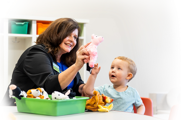 Smiling teacher holds up a pink pig puppet while interacting with a young child during a playful learning activity. The child looks engaged and curious, surrounded by a bin of stuffed animals on the table.