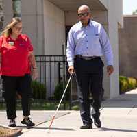Man who is blind walking outside Envision Main Street building with white cane and an Envision employee