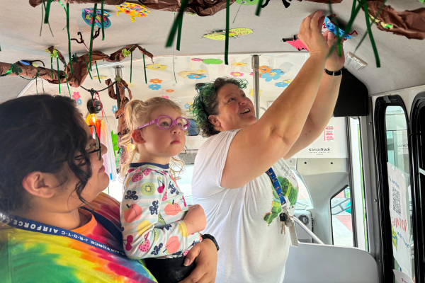 An adult helps a young child decorate the ceiling of a bus with colorful hanging artwork.
