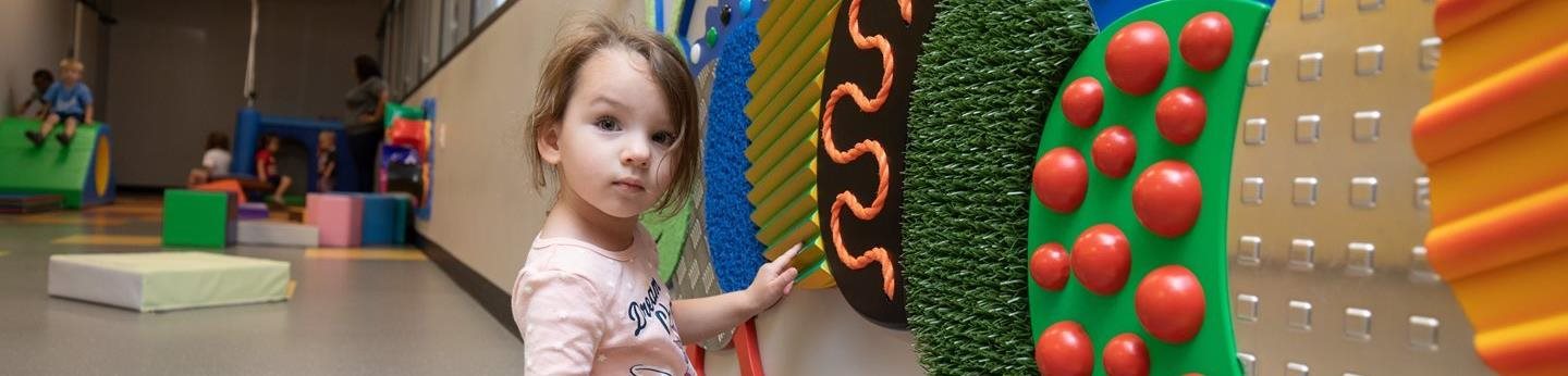 Young child interacting with a colorful sensory wall at the Envision Child Development Center in Wichita, KS. The indoor play area features a variety of tactile textures and shapes to support explorat