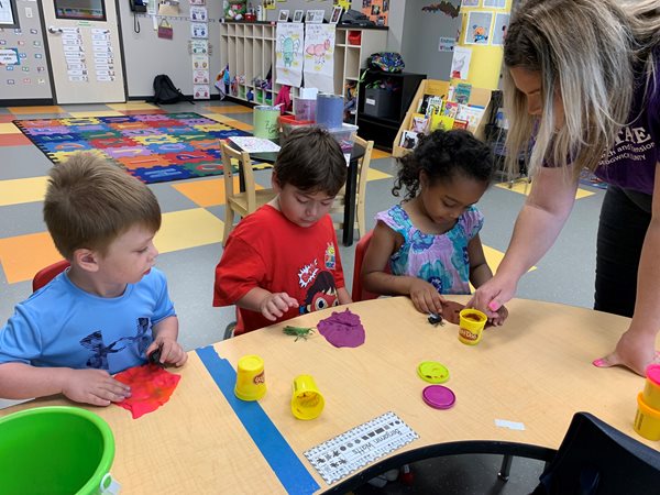 Three children are sitting at a wooden table working with playdough as the teacher helps them.