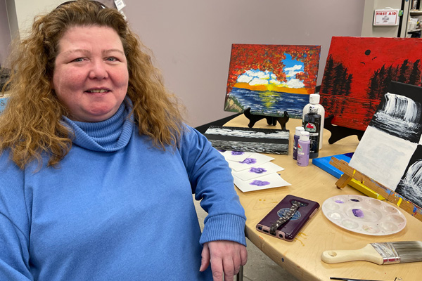A woman in a blue shirt sitting at a wooden desk smiling for a photo with paintings behind her