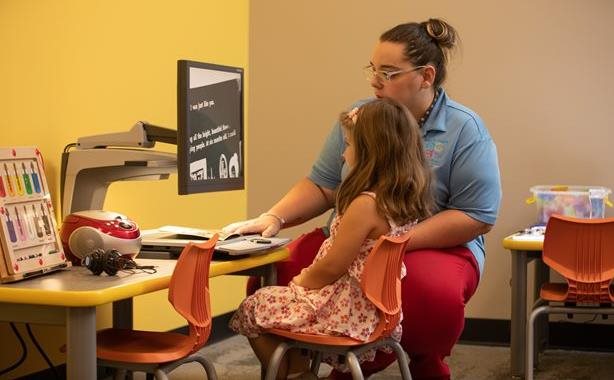 A teacher works one-on-one with a young girl in a classroom, using an accessible video magnifier to help her read printed material. The setting includes colorful chairs and educational tools designed to support children with visual impairments.