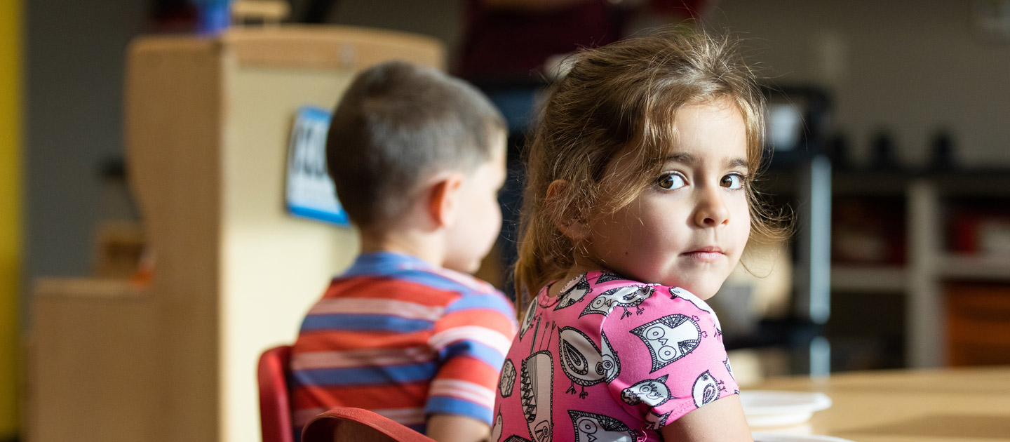 Little girl with big brown eyes in a pink dress sits with her friend at a table in ECDC at Envision.