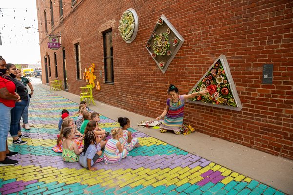 Tomiyo sit in front of a brick wall in Gallery Alley in front of her geometric and colorful ceramic flower art piece with a group of students from Envision Child Development Center