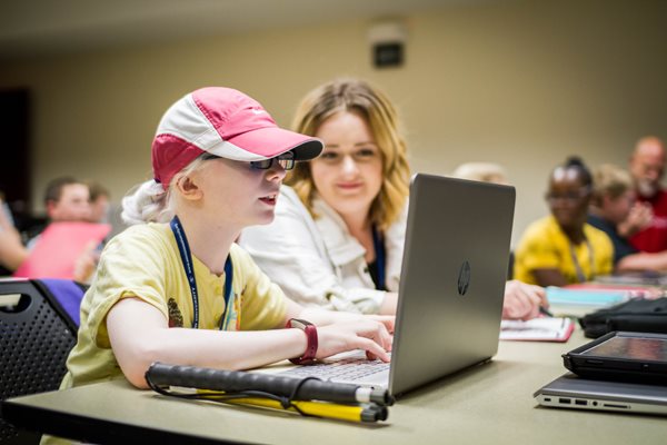 A girl wearing a pink and white hat working on a laptop computer with her mentor sitting beside her.