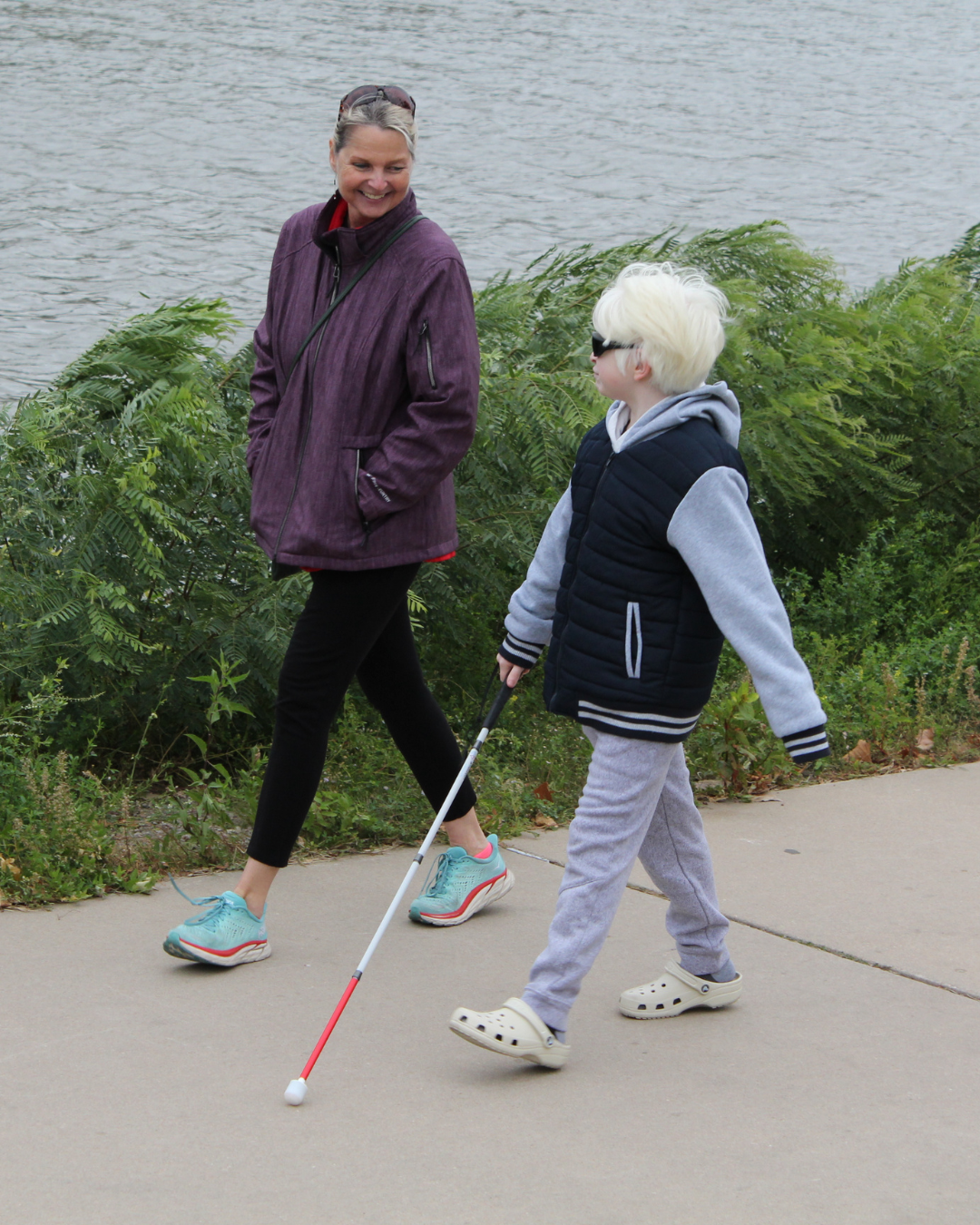 a young boy walking with a white cane as his mom walking next to him smiles.