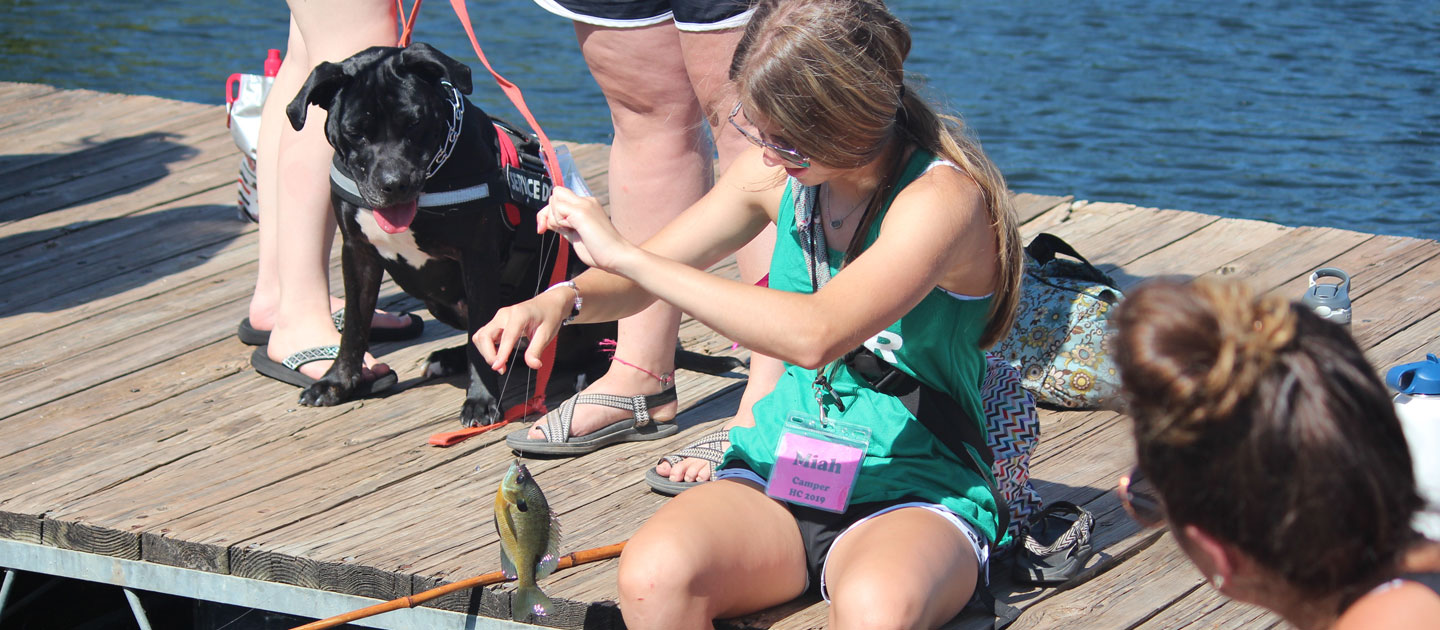 Girl catches a fish at the lake while a guide dog looks on at Heathers Camp.