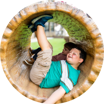 Two young boys playing inside a large, hollow log-shaped tunnel at an outdoor playground. One boy is lying on his back with his legs up, while the other leans against the inside of the tunnel, both appearing to enjoy the playful moment.