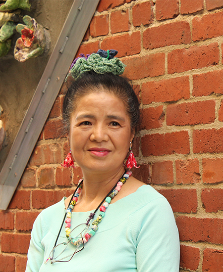 Tomiyo stands in front of a brick wall in Gallery Alley in front of her geometric and colorful ceramic flower art piece
