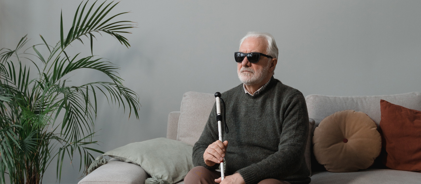 Older man who is blind sits on a couch in his living room holding his white cane.